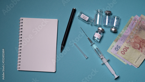 Blank notebook with pen on a blue background. Empty vaccine vials and a syringe with a needle lie on the table. Concept: Vaccine, injection, money, counting.