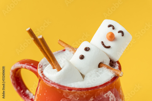Marshmallow snowman taking hot tub in a red ceramic cup full of cocoa with milk foam. Christmas holidays yellow background. Wintertime concept. Hot chocolate with marshmallow and festive decoration.