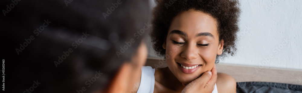lesbian african american woman touching cheek of happy girlfriend in bedroom, banner. lesbian african american woman touching cheek of happy girlfriend in bedroom, banner.