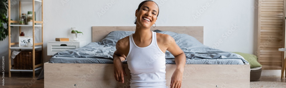 young and joyful african american woman in tank top sitting near bed, banner.