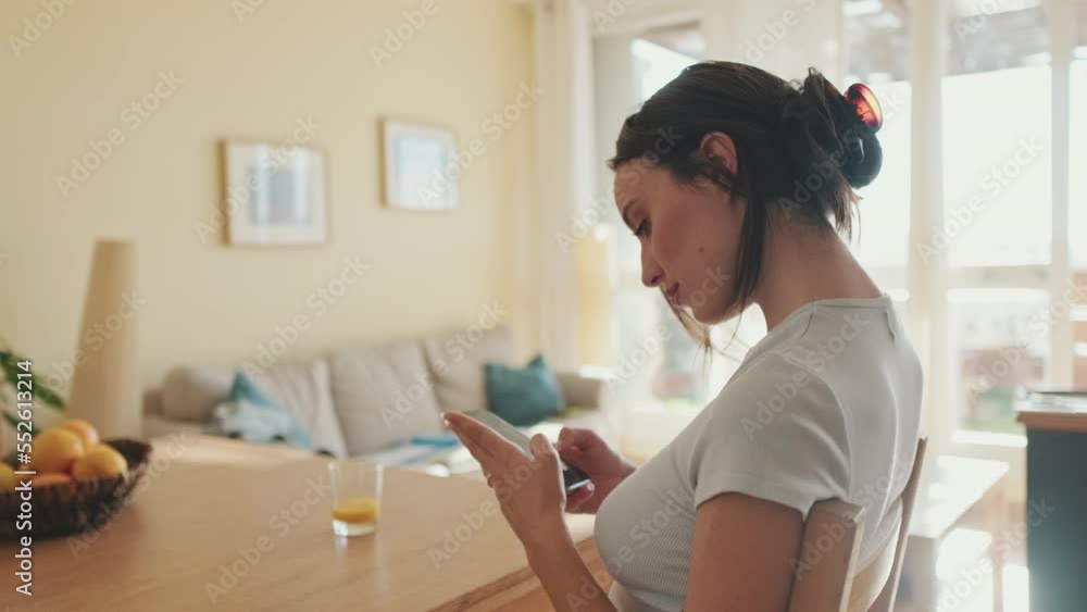 Smiling young woman scrolling through photo on her mobile phone while sitting in the kitchen Smiling young woman scrolling through photo on her mobile phone while sitting in the kitchen