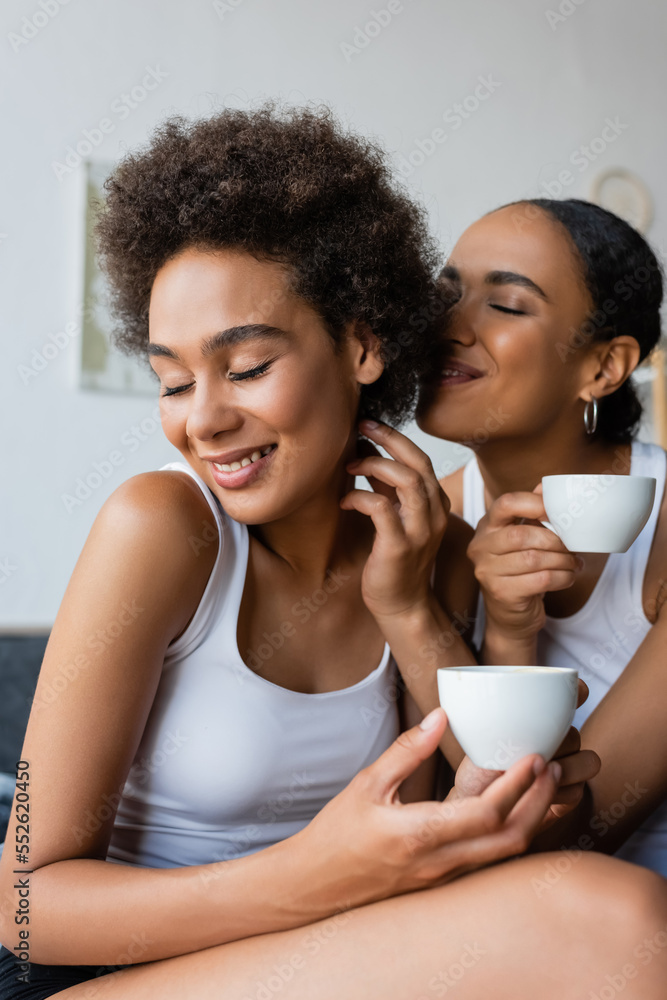 happy lesbian african american couple holding cups of coffee while smiling at home. happy lesbian african american couple holding cups of coffee while smiling at home.