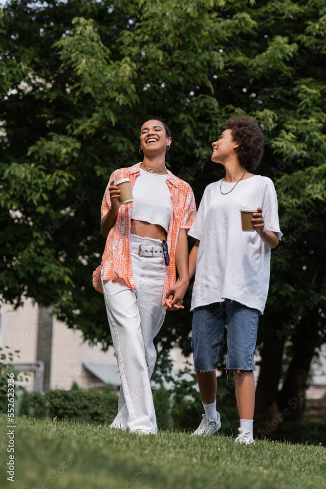 full length of joyful african american lesbian women holding paper cups and standing in park.