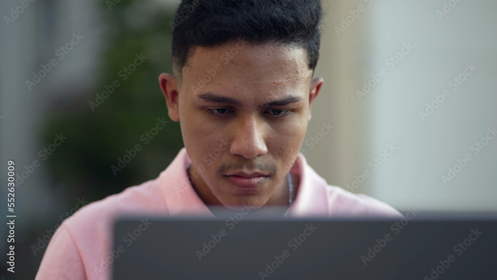 One young hispanic man closeup face looking at computer laptop outside One young hispanic man closeup face looking at computer laptop outside