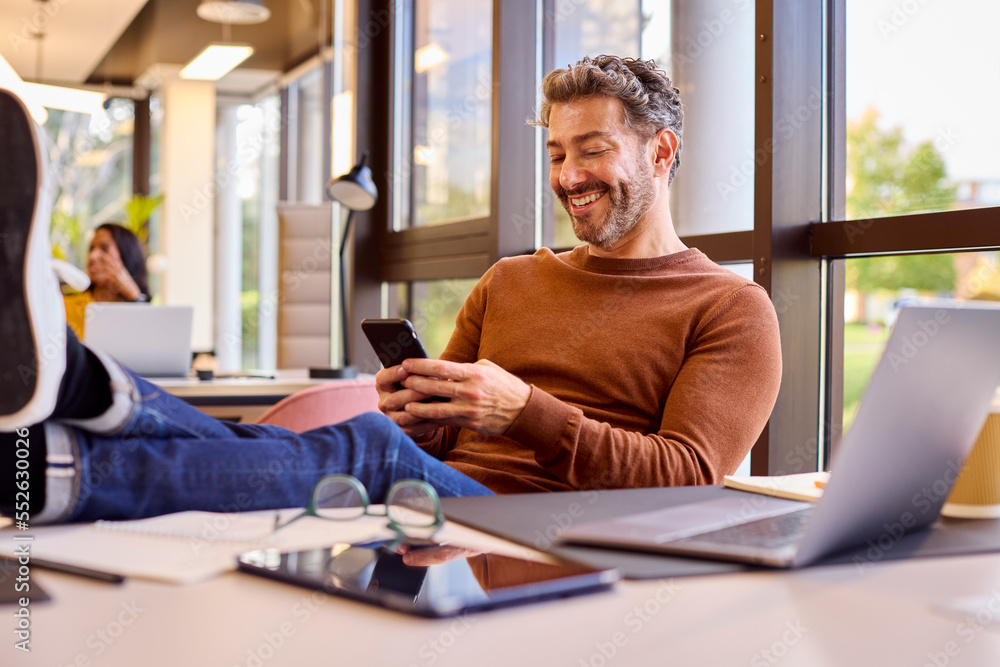 Obraz premium Casually Dressed Mature Businessman With Feet On Desk In Office Using Mobile Phone