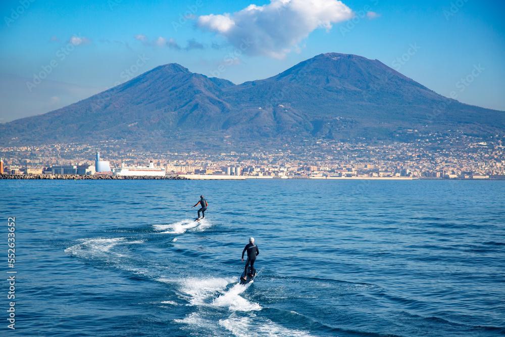 Two people ride an electric surfboard in the Gulf of Naples, Italy, against the backdrop of Mount Vesuvius