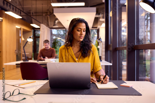 Free Download Mature Businesswoman Working On Laptop At Desk In Office Writing In Notebook - GFXLoad