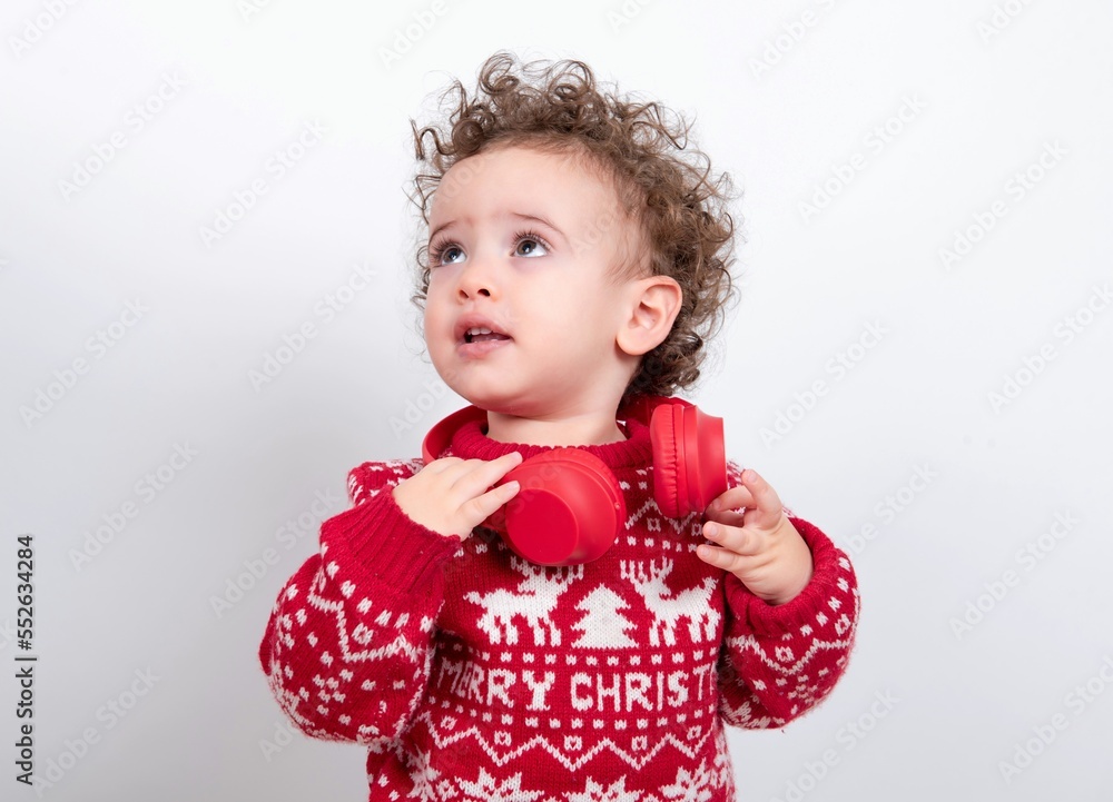 Beautiful little baby boy with curly hair wearing Christmas knitted sweater against white background wearing red headphones listening to music and looking aside. 