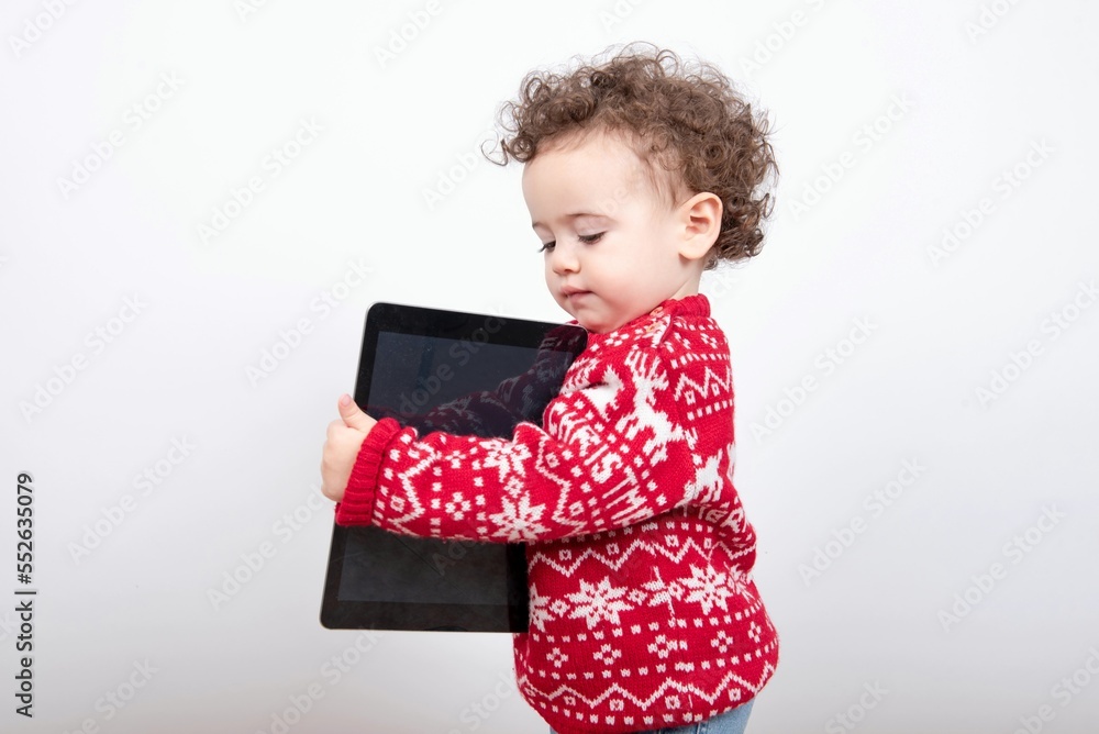 Beautiful little baby boy with curly hair wearing red Christmas knitted sweater against white background holding tablet pad for watching streaming videos and playing educational video games. Beautiful little baby boy with curly hair wearing red Christmas knitted sweater against white background holding tablet pad for watching streaming videos and playing educational video games.
