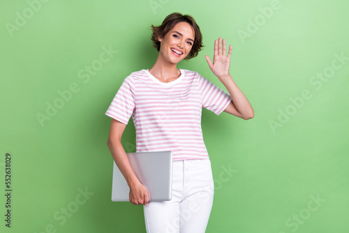 Portrait photo of young adorable pretty smiling woman hold laptop from work showing high five palm hello employer isolated on green color background