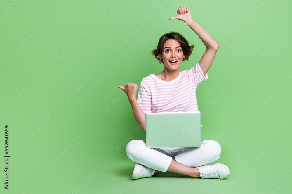 Full size photo of pretty young lady folded legs netbook point empty space dressed trendy striped outfit isolated on green color background Full size photo of pretty young lady folded legs netbook point empty space dressed trendy striped outfit isolated on green color background