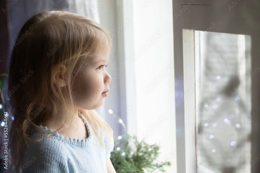 bored cute little girl looking out of window on background sparkle blurred lights at home