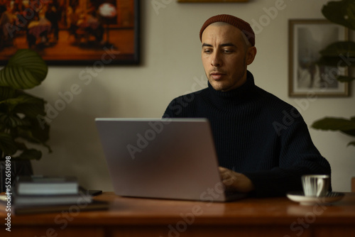 A caucasian man wearing a beanie and a knitted sweatshirt sitting at a wooden table working on a laptop.