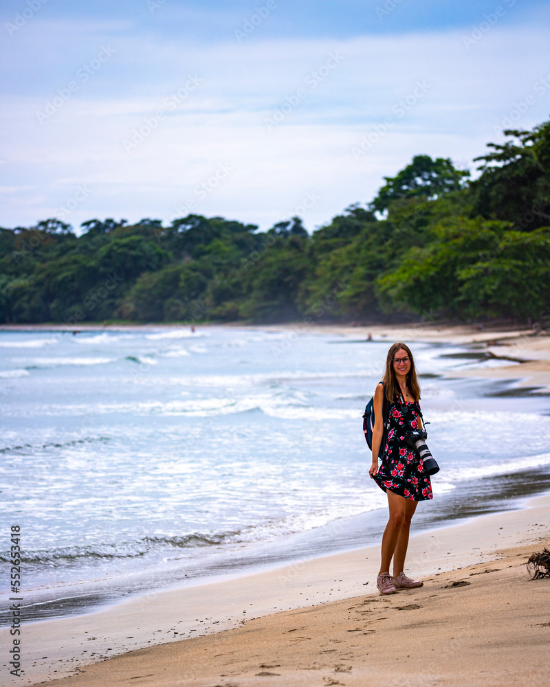 A beautiful backpacker girl with a camera walks on a tropical beach on the Caribbean coast of Costa Rica; beach vacation in Costa Rica