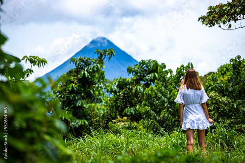 A beautiful girl in a white dress admires the view of a volcano arenal in Costa Rica; a massive volcano seen from a tropical rainforest photo