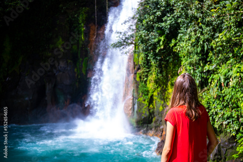 A BEAUTIFUL GIRL IN A RED DRESS STANDS UNDER THE FAMOUS BLUE WATERFALL OF RIO CELESTE, COSTA RICA; A WATERFALL WITH BLUE WATER IN VOLCANO TENORIO NATIONAL PARK; A WATERFALL IN THE RAINFOREST