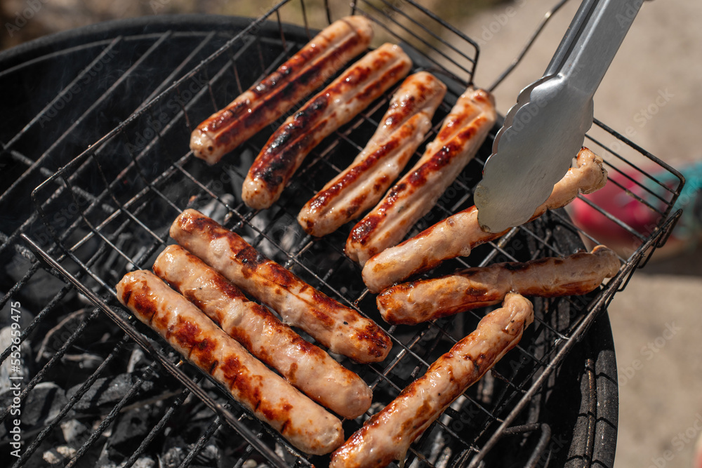 Closeup photo of sausages. Grilling chicken sausages outside on a barbecue grill. Grill tongs in the foreground. First outdoor grill party in the spring season. Healthy barbecue. 