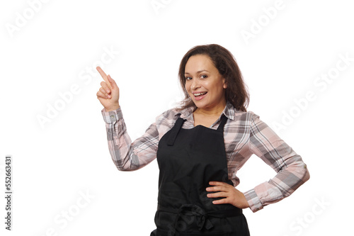 Multi-ethnic positive cheerful pretty woman barmaid, waitress, bartender in black chef apron, holding a copy space for your advertising text on her hand palm up, smiles at camera over white background