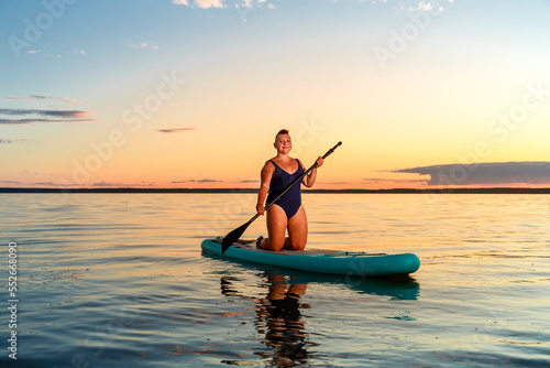 a Jewish feminist woman in a closed bathing suit with a mohawk on a koyen on a stand-up board with an oar floats on the water against the background of the sunset sky.