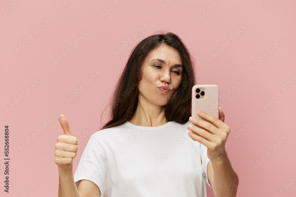 beautiful cute woman with a trendy phone in her hands stands on a pink background in a white t-shirt, holds her thumb up. Horizontal photo on a pink background beautiful cute woman with a trendy phone in her hands stands on a pink background in a white t-shirt, holds her thumb up. Horizontal photo on a pink background
