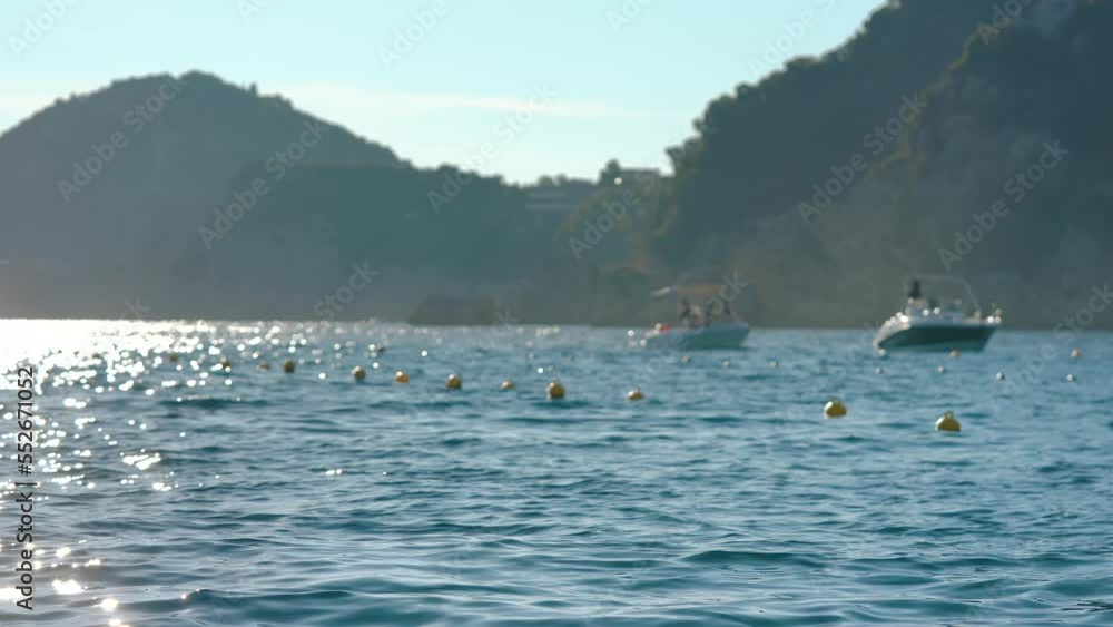 View of Liapades bay in Greece, focus on rather calm sea with small waves in foreground, blurred boats moving slowly with rocky cliffs background. Afternoon sunlight haze scene