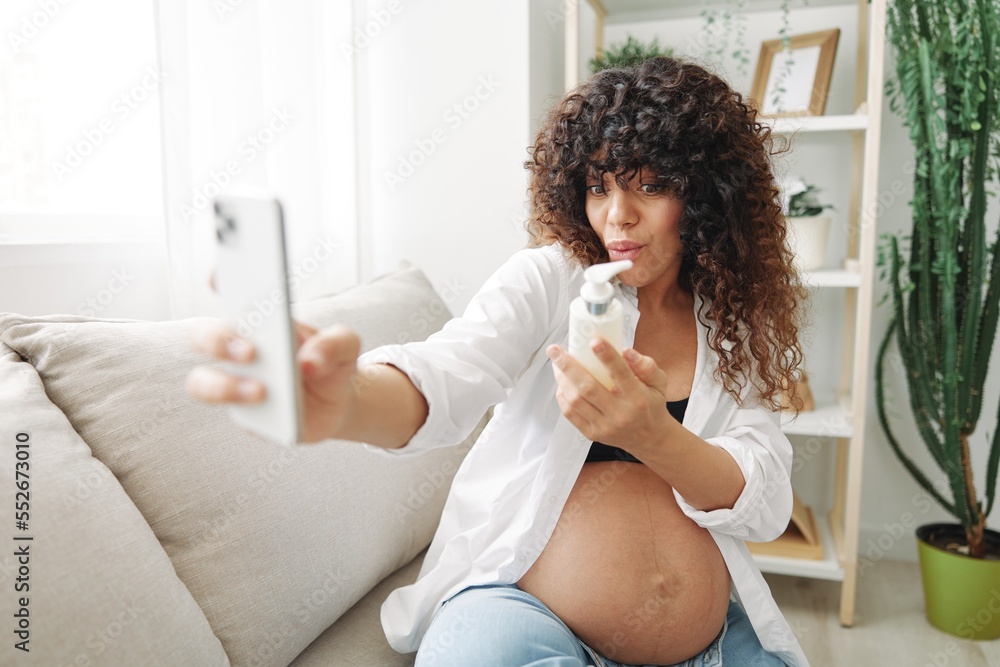 Pregnant woman smile blogger cream for pregnant women stretch marks body during pregnancy filming herself on phone, video chat sitting on the couch at home freelancer in the last month of pregnancy Pregnant woman smile blogger cream for pregnant women stretch marks body during pregnancy filming herself on phone, video chat sitting on the couch at home freelancer in the last month of pregnancy