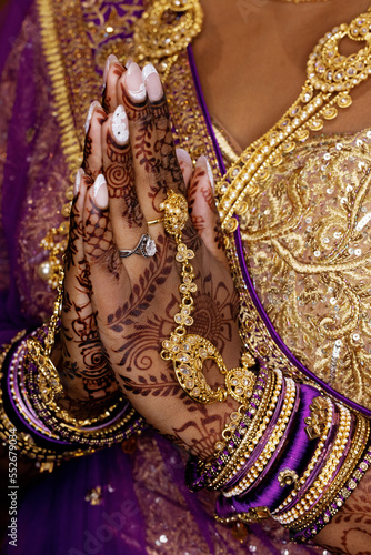 Close-up hands of woman bride in a traditional wedding purple dress, having golden jewelry on her fingers, and henna drawings holding hands together and praying