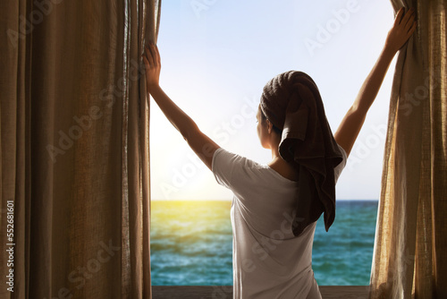 The girl from the back opens the curtains. Girl with a towel on her head. View from the window to the sea © MM