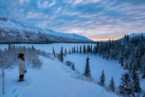 Woman standing in the snow on a mountain top looking out at a stunning landscape with the sun rising over Annie Lake in Winter; Whitehorse, Yukon, Canada