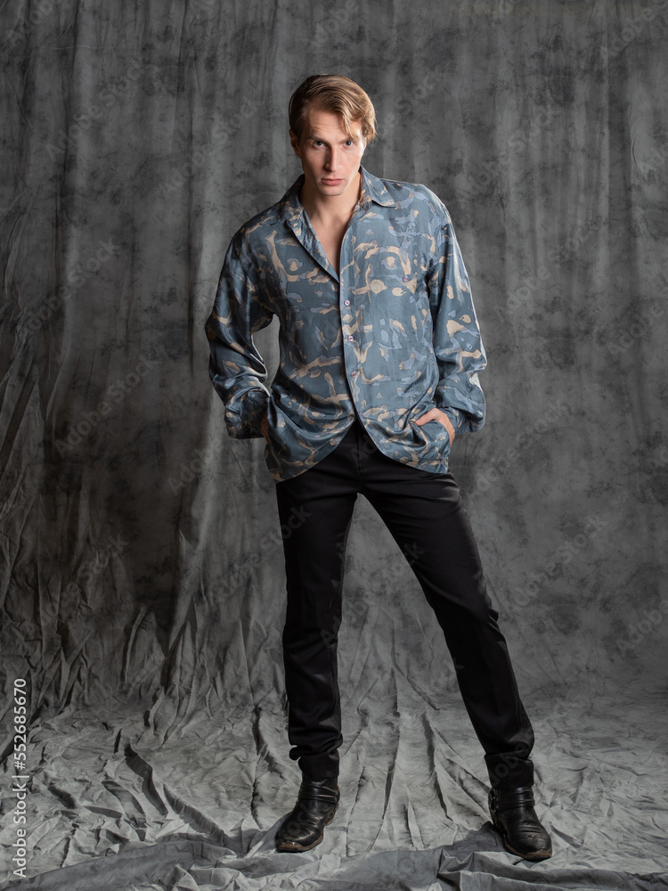Stylish elegant young man in a blue silk shirt posing in the studio on a gray fabric background Stylish elegant young man in a blue silk shirt posing in the studio on a gray fabric background