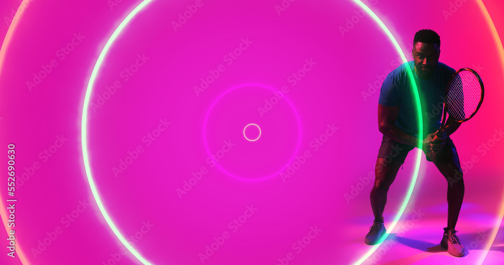 African american male player holding racket and standing over illuminated circles on pink background African american male player holding racket and standing over illuminated circles on pink background