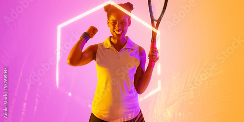 African american female tennis player holding racket and shaking fist by glowing hexagon and plants