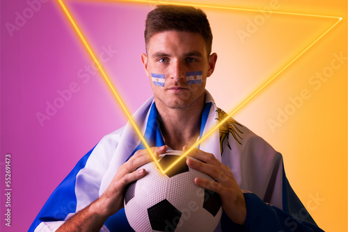 Male caucasian fan holding soccer ball with flag of argentina and face paint by illuminated triangle