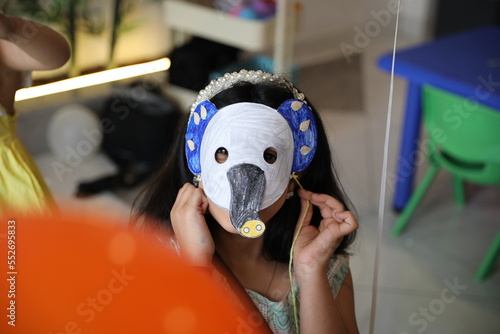 A little child girl wearing a colored animal paper mask fronting her face is isolated in the classroom background with an orange balloon. elephant paper mask art and crafts.