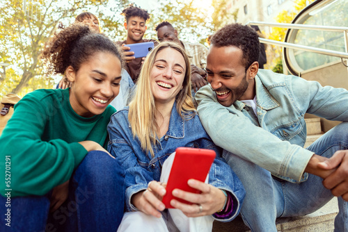 United group of international friends laughing together watching social media content on mobile phone app. Diverse millennial young adult people laughing together while taking vertical selfie portrait