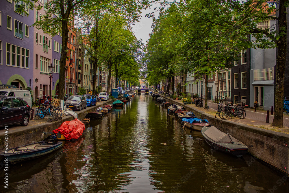 A canal in Amsterdam, boats on the canal, parked cars, trees and historic buildings.