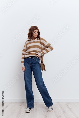 a full-length photo of a beautiful, stylish woman standing on a light background in a striped sweater and blue jeans, posing relaxed with a beige bag slung over her shoulder