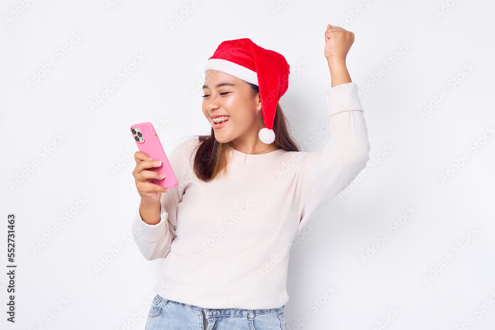 Excited young Asian woman in a Christmas hat using smartphone and doing winner gesture isolated over white background. celebration Christmas holiday and New Year concept Excited young Asian woman in a Christmas hat using smartphone and doing winner gesture isolated over white background. celebration Christmas holiday and New Year concept