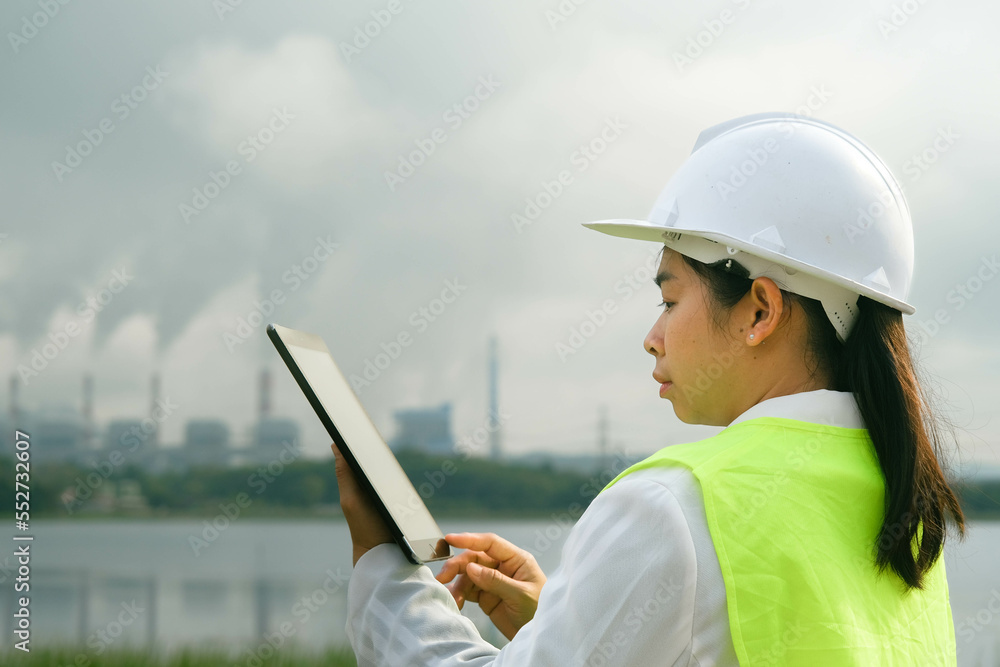 Female chief engineer wearing a green vest and helmet stands outside against the background of coal power plant station and steam in the morning mist. Engineer working and laptop at coal power plant. Female chief engineer wearing a green vest and helmet stands outside against the background of coal power plant station and steam in the morning mist. Engineer working and laptop at coal power plant.