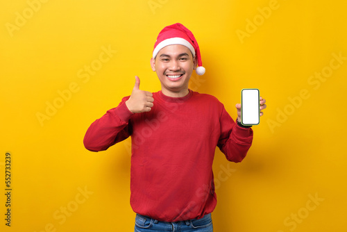 Cheerful young Asian man in Santa hat holding mobile phone with blank screen, showing thumb up gesture over yellow studio background. celebration Christmas holiday and New Year concept © Sewupari Studio