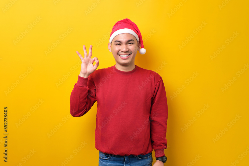 Cheerful young Asian man in Santa hat showing ok sign and looking at camera over yellow studio background. celebration Christmas holiday and New Year concept Cheerful young Asian man in Santa hat showing ok sign and looking at camera over yellow studio background. celebration Christmas holiday and New Year concept