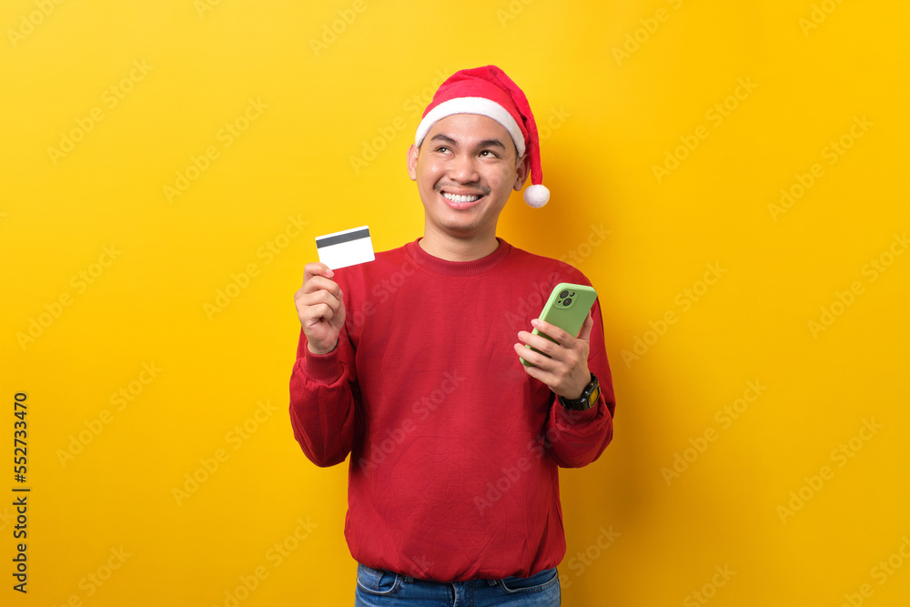 Smiling young Asian man in Santa hat using mobile phone and credit card, looking up, over yellow studio background. celebration Christmas holiday and New Year concept Smiling young Asian man in Santa hat using mobile phone and credit card, looking up, over yellow studio background. celebration Christmas holiday and New Year concept