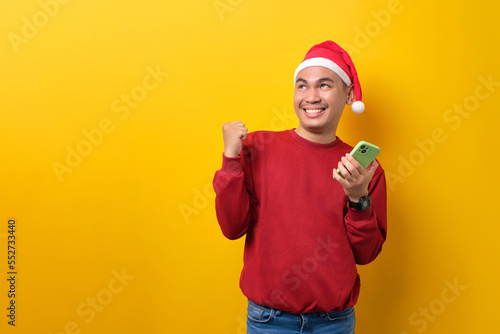 Excited young Asian man in Santa hat holding mobile phone, raising hands up, looking away at copy space over yellow studio background. celebration Christmas holiday and New Year concept © Sewupari Studio