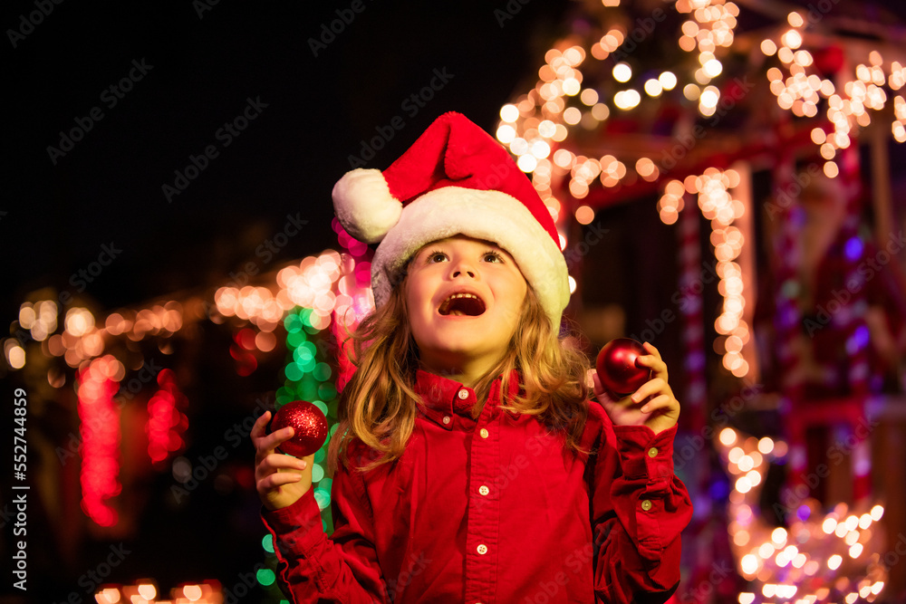 Christmas decoration in front of a night house. Happy kid looks at the sky in the Christmas night. Kid in santa hat near home garlands outside. House Decorated with Garlands. Christmas decoration in front of a night house. Happy kid looks at the sky in the Christmas night. Kid in santa hat near home garlands outside. House Decorated with Garlands.