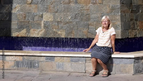 Blond woman sitting and enjoying the sun while looking around by a small artificial waterfall outside La Zenia shopping center in Torrevieja, Spain photo