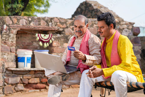 Young indian farmer using card and showing some detail to his father in laptop.