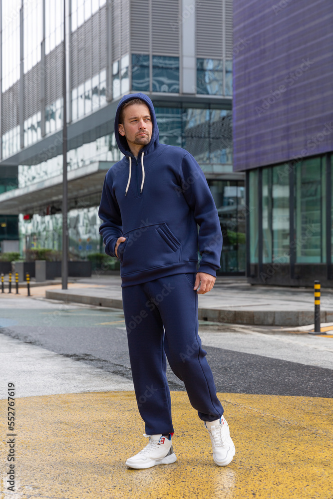 Full-length young man wears a blue sports suit and poses for the camera. Athletic handsome young man stands on urban location and shows sports suit. Mock-up Full-length young man wears a blue sports suit and poses for the camera. Athletic handsome young man stands on urban location and shows sports suit. Mock-up