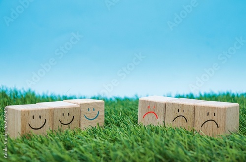 sad faces icon and happy smiley faces on wooden cube blocks on grass and blue background. concept of positivism, mental well-being.