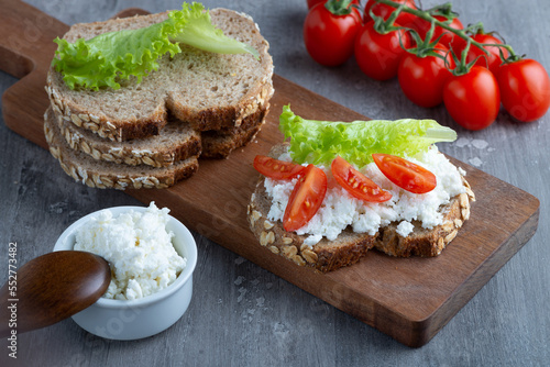 Slices of rye bread with cottage cheese and tomatoes on wooden plate on grey background