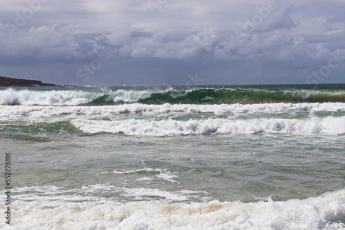 Waves crashing at Fingal Beach with people walking along the sand in the distance on a sunny day.